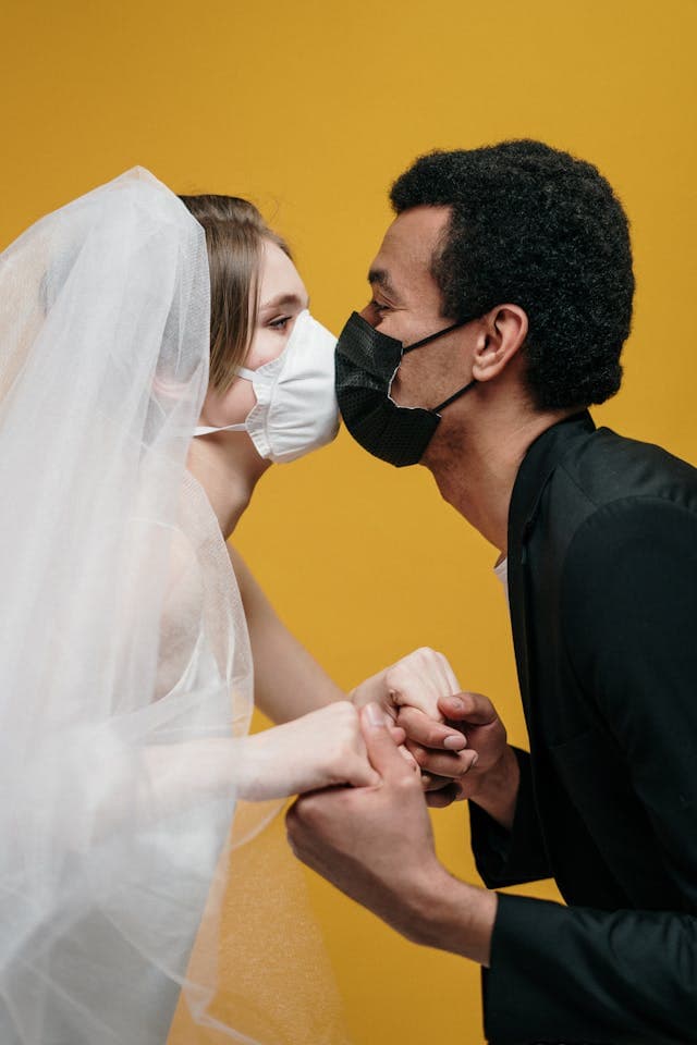 man in black suit jacket holding his bride in white wedding dress and kissing with mask on signifying the importance of vaccines before the wedding to prevent diseases